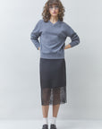 A model stands against a light gray studio background wearing a heathered blue-gray V-neck sweater, a black midi skirt with a sheer black lace hem, white socks, and black loafers.