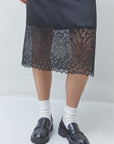 A person wearing a black midi skirt with a sheer floral lace hem, paired with white socks and black leather loafers, shown from the waist down against a light gray studio background.