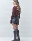 A woman is shown from the back wearing a burgundy long-sleeve top, black leather shorts, gray socks, and black knee-high boots against a light gray studio background.
