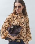 A woman wearing a brown and beige animal-print blouse, dark sunglasses, and a dark brown handbag, standing against a light gray studio background.