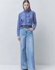 A model stands against a light gray studio background wearing a blue and white gingham long-sleeve blouse with a high ruffled mock neck, paired with wide-leg light blue jeans and a dark belt.