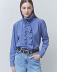 A woman modeling a blue and white gingham long-sleeve blouse with a high ruffled mock neck, paired with light-wash blue jeans and a brown belt, against a solid light gray background.