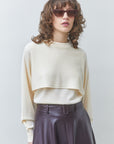 A model with curly brown hair and sunglasses wearing a cream-colored cropped long-sleeve knit top and a dark purple high-waisted leather skirt, set against a plain light gray studio background.
