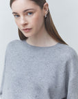 A woman wearing a light gray knit sweater adorned with small silver rhinestones, photographed against a plain light gray background.