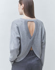 A woman shown from the back wearing a light gray knit sweater with a large open V-back design and dark gray trousers, photographed against a plain light gray studio background.