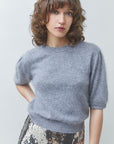 A woman with shoulder-length brown hair wearing a light gray short-sleeve knit sweater and a patterned metallic skirt, photographed against a plain light gray studio background.