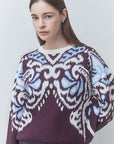 A woman wearing a dark purple knit sweater with a white and light blue ornate pattern across the chest and sleeves, paired against a light gray studio background.