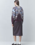 A model is shown from the back wearing a dark purple knit sweater with a white and light blue ornate pattern, paired with a dark purple midi skirt, white socks, and black loafers, against a light gray studio background.