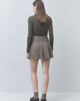 A model shown from the back wearing an olive green ribbed long-sleeve top and a brown and gray plaid pleated mini skirt, paired with gray socks and black platform shoes, against a light gray studio background.