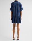 A model viewed from the back wearing a short-sleeved blue tiered shirt dress with a collar and pleated back detail, paired with black heels, against a light gray studio background.