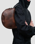 A person wearing a black hooded jacket carries a large, semi-circular brown leather shoulder bag against their back, shown against a plain light gray background.
