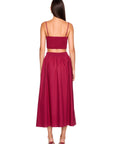 A woman viewed from the back wearing a matching two-piece outfit consisting of a deep magenta spaghetti-strap crop top and a high-waisted pleated midi skirt, paired with dark brown heels against a plain white background.