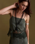 A woman wearing a dark olive green camisole dress with lace trim and a long black beaded tassel necklace, posed against a solid olive-brown background.
