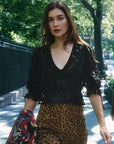 A woman walking outdoors on a tree-lined sidewalk wearing a black ruffled lace blouse and leopard-print pants, holding a floral garment in one hand.