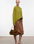 A model stands in a minimalist studio setting wearing a loose olive green knit sweater, a brown satin midi skirt, and dark brown pointed-toe mules, while carrying a brown suede flap shoulder bag.