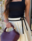 Woman holding a purple handbag wearing a black top and white skirt with a black belt.
