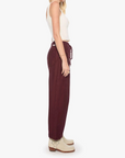 A woman shown in profile wearing loose, wide-leg burgundy drawstring pants, a white ribbed tank top, and cream ankle boots against a plain light gray studio background.