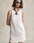 Woman wearing a white sleeveless dress with sunglasses and a necklace against a neutral background