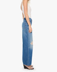 A woman shown in profile wearing a white sleeveless top and high-waisted, wide-leg blue denim jeans with a distressed knee detail, set against a plain light gray background.