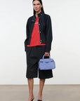 A model stands in a studio wearing a black cropped jacket over a red graphic T-shirt, black shorts, and black heeled sandals, while holding a light blue structured handbag.