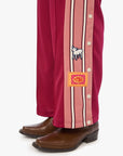 Close-up of a person's legs wearing pink and burgundy striped pants with decorative patches and brown leather boots, photographed against a clean white background.