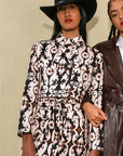 Two women standing side by side, one wearing a patterned outfit and black hat, the other in a brown coat.
