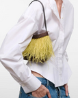 A model wearing a white button-down shirt and blue jeans, carrying a small yellow fringe handbag with a dark brown top band and black strap against a plain white studio background.