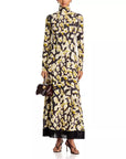 A model wears a long-sleeved high-neck maxi dress featuring a black base with a yellow and cream floral print and a black fringe hem. She is holding a small dark brown handbag and wearing black strappy heels against a plain white background.