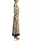 A long-sleeved maxi dress with a high mock neck, featuring an all-over floral print in cream and olive green on a black background, with black fringe detailing along the hem. The dress is shown from the back on a model against a white studio background, side view