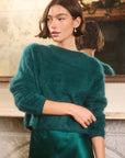 A woman wearing a fuzzy teal-green knit sweater and a satin green skirt, standing indoors in front of a mirror that reflects her back.