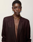 A model with short dark hair is wearing a dark brown blazer, a chunky gold chain necklace, and Luminary Earrings, posed against a solid light beige background.
