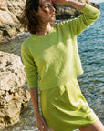 A woman standing on a rocky shoreline by the sea, wearing a bright lime green knit sweater and matching mini skirt.