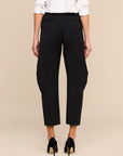 Rear view of a model wearing black cropped trousers with side pockets, paired with a white blouse and black heels, against a solid beige background.