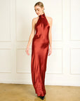 Woman wearing a red satin halter neck gown against a white paneled wall, front