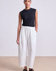 Woman wearing a black sleeveless top and white pants against a plain background