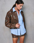 Woman wearing a brown embroidered jacket over a light blue shirt against a gray background, front