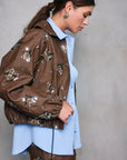 Woman wearing a brown jacket with floral embellishments against a plain background, side view