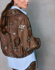 Woman wearing a brown jacket with floral embellishments against a gray background
