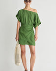 Woman wearing a green off-shoulder dress holding a beige woven bag on a white background