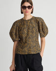 A model wearing a short-sleeved puff-sleeve blouse with a brown and black leopard print, paired with black trousers and sunglasses, against a plain light gray background.