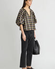 model wearing a plaid vneck short sleeve top with black jeans carrying a black bag on a grey background, side view