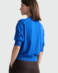 A woman shown from the back wearing a bright blue knit sweater with short puff sleeves and dark brown trousers, photographed against a clean white studio background.