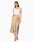 A woman modeling a champagne beige satin midi skirt with a lace side panel and hem, paired with a white crop top and white sandals, against a plain white background.