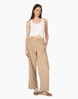A model stands against a plain white background wearing beige wide-leg trousers, a white cropped tank top, and brown sneakers.