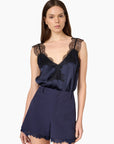 A woman modeling a navy blue satin camisole top with black lace trim and matching navy shorts with lace hem, against a plain white background.