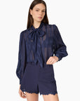 A model wearing a navy blue sheer long-sleeve blouse with a large bow tie-neck and matching navy blue shorts with a scalloped lace hem, set against a plain white studio background.