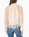 A woman viewed from the back wearing a sheer cream lace blouse with long sleeves and a gathered neckline, paired with blue jeans against a plain light background.