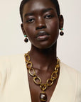A close-up portrait of a model wearing a chunky gold chain necklace with a dark brown pendant, Umbria Earrings in Tigers Eye, and a cream-colored blazer against a light beige background.