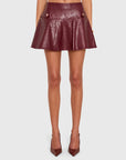 A model from the waist down wearing a high-waisted, flared faux-leather mini skirt in a deep burgundy color, paired with matching pointed-toe heels, against a light gray background.