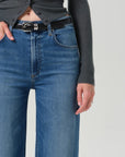 Close-up of a person wearing blue denim jeans with a black belt, paired with a dark gray ribbed top, against a light gray studio background.